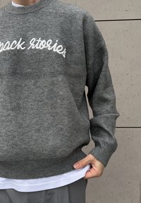Grauer Strickpullover mit gerippten Bündchen und Saum, mit dem bestickten Schriftzug "pack stories" in Weiß über der Brust.