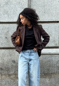 Młoda kobieta w kręconych włosach w brązowej marynarce, czarnej bluzce, jasnoniebieskich dżinsach i brązowej torebce, stoi przed szarym kamiennym murem.