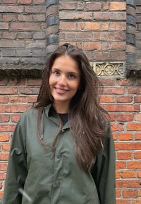 Giacca impermeabile verde oliva con chiusura frontale a bottoni, indossata sopra una maglia nera. Capelli lunghi e scuri, fiocchi di neve leggeri visibili sulla giacca. Sfondo con muro di mattoni.