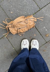 Svetlo rjava tkaninska torba z zadrgami in resicami na sivi asfaltni površini, v kombinaciji z belimi in sivimi športnimi čevlji ter modrimi jeans hlačami.