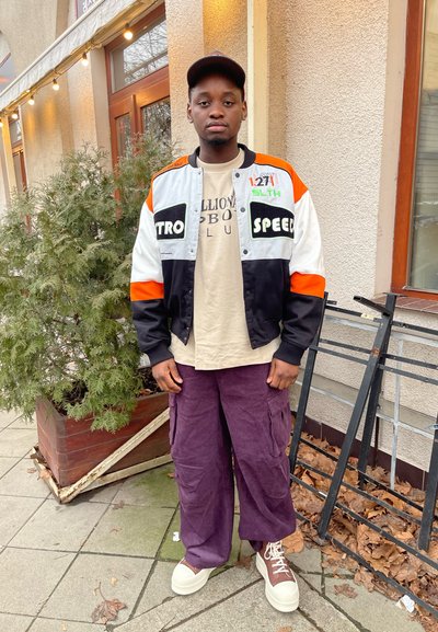 Chaqueta varsity negra y naranja sobre una camisa beige, combinada con pantalones cargo morados y zapatillas crema con detalles marrones.