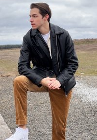Svart konstläderjacka med dragkedja, tillsammans med beige kordbyxor. Modellen står på ett ben, iförd vita sneakers.