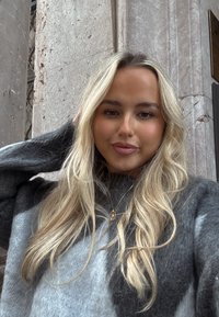 Jeune femme aux longs cheveux blonds portant un pull gris et noir, debout contre un mur en pierre texturé, regardant la caméra.