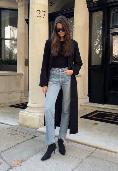 Abrigo largo negro sobre una blusa negra, jeans de mezclilla azul claro y tacones negros con calcetines, de pie en un camino de piedra con un fondo de un edificio alto.