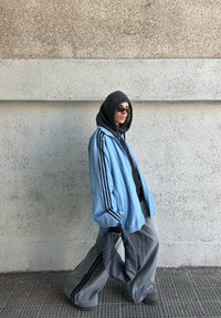 Personne portant une veste bleu clair oversize avec des bandes noires, un pantalon large gris, un sweat à capuche noir, des lunettes de soleil, et tenant un sac à motif noir et blanc en marchant.
