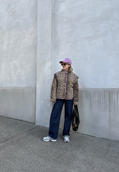 Chaqueta de leopardo con detalles morados, combinada con jeans de pierna ancha de color azul oscuro y zapatillas blancas, complementada con una gorra morada y un bolso negro.