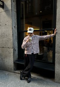 Chemise à carreaux en tissu doux, pantalon noir ample et chaussures noires. Tient un verre avec une paille, debout contre une fenêtre de café.