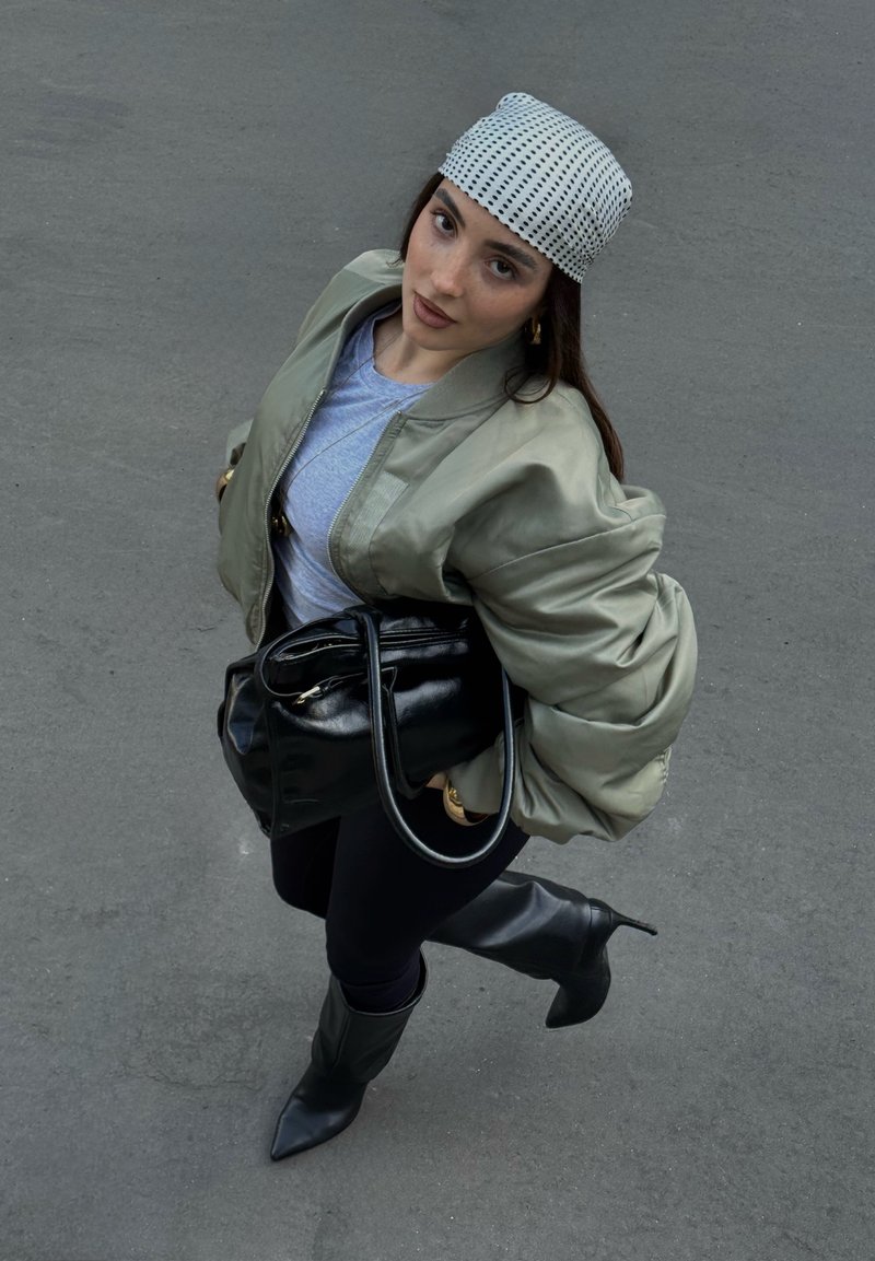 Olive green bomber jacket, gray t-shirt, black handbag, black leggings, knee-high pointed boots, and a white checkered headscarf.