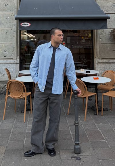 Camisa de botones azul clara, camiseta negra, pantalones de pierna ancha grises y zapatos negros. Escenario de café al aire libre con mesas redondas y sillas de mimbre.