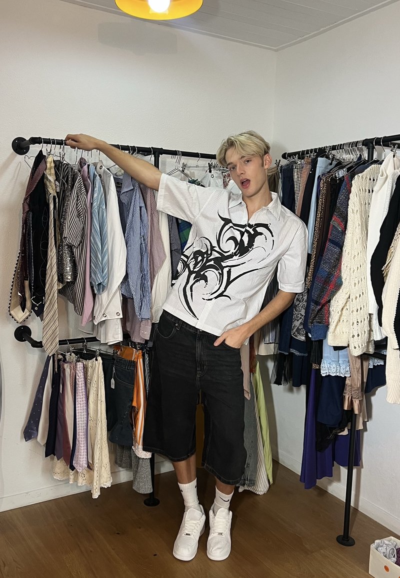 Jeune homme portant un short noir et un t-shirt blanc à motif, se tenant dans une pièce en tenant un portant à vêtements rempli de chemises, cravates et pulls.