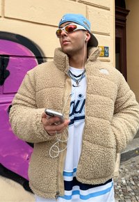 Beige φλις μπουφάν με υφή και πλούσια εξωτερική πλευρά, με κουκούλα, φορεμένο πάνω από ένα λευκό μπλουζάκι με μπλε λεπτομέρειες, κρατώντας ένα smartphone.