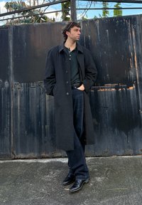 Άνδρας με μαύρο μακρύ παλτό, μαύρο πουκάμισο, τζιν και μποτάκια στέκεται με τα χέρια στις τσέπες μπροστά σε μια ψηλή μαύρη μεταλλική πόρτα.