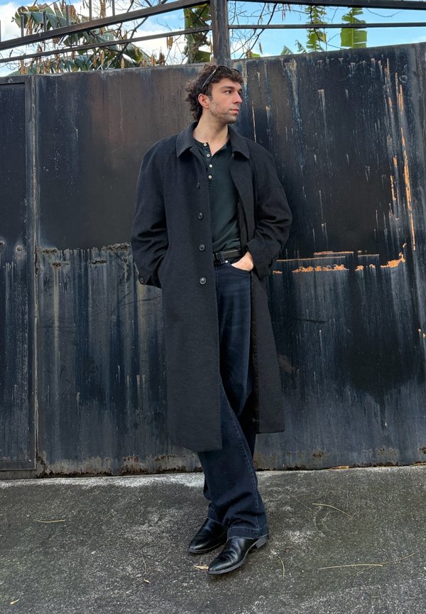 Uomo con lungo cappotto nero, camicia nera, jeans e stivali, in piedi con le mani nelle tasche davanti a un alto cancello di metallo nero.