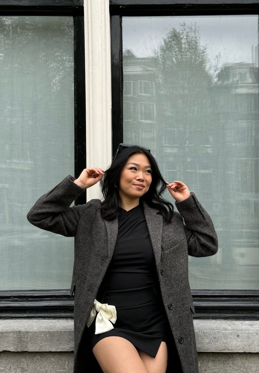 Femme portant un manteau en laine gris foncé et une robe noire avec un accent de nœud blanc. Elle pose devant une fenêtre avec des rideaux verts.