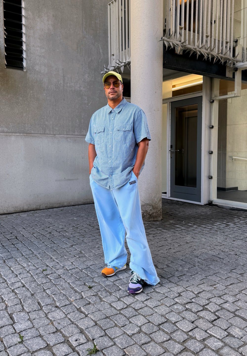 Svetlo modri široki hlače, denim srajca s kratkimi rokavi, rumena kapica, večbarvni čevlji, stojijo ob betonski steni.