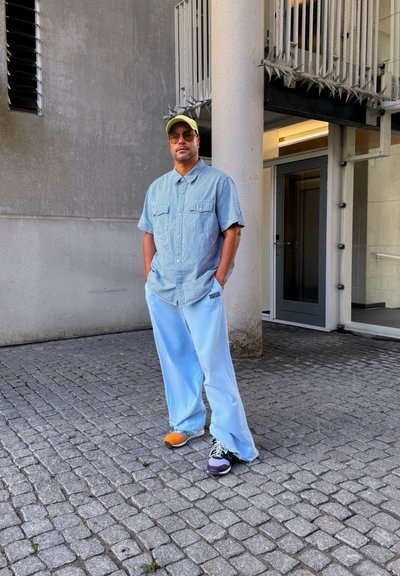 Pantalones oversize de color azul claro, camisa de denim de manga corta, gorra amarilla, zapatos multicolores, de pie contra una pared de concreto.