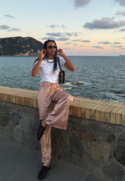 Pantalones anchos de satén rosa claro, camiseta blanca, zapatos de slip-on negros trenzados, collar de cadena dorado, de pie sobre un muro de piedra junto al mar.