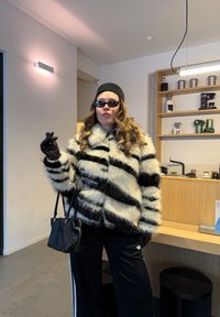 Femme portant un manteau en fausse fourrure à rayures noires et blanches, des gants noirs, des lunettes de soleil et un chapeau, tenant un sac à main noir dans une cuisine moderne.