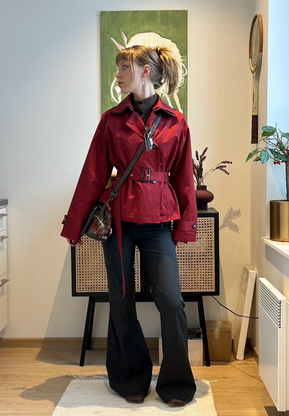 Personne portant une veste rouge ceinturée, un pantalon noir évasé et un sac bandoulière, debout à l'intérieur avec des peintures et des plantes en arrière-plan.