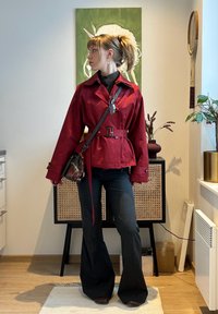 Personne portant une veste rouge ceinturée, un pantalon noir évasé et un sac bandoulière, debout à l'intérieur avec des peintures et des plantes en arrière-plan.