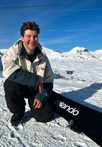 Joven con chaqueta de invierno arrodillado sobre la nieve junto a una tabla de snowboard con el logo de "zalando", montañas nevadas y cielo despejado al fondo.