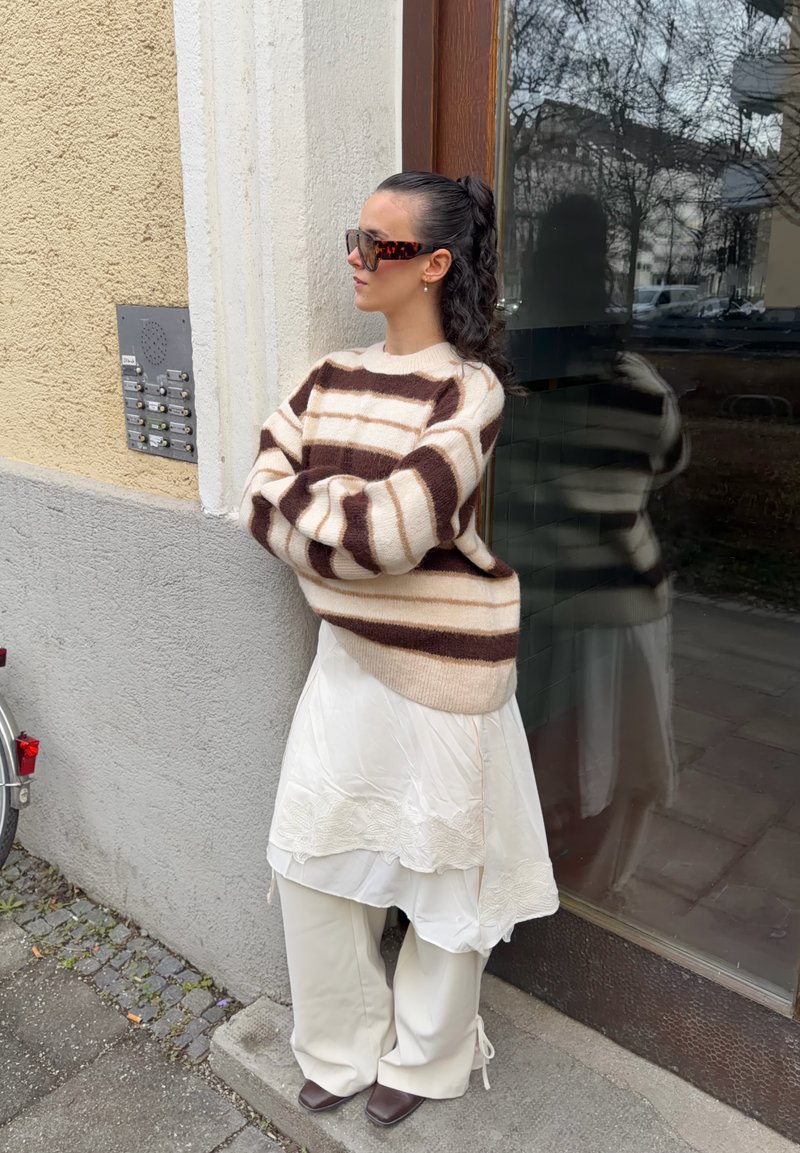 Femme aux cheveux foncés attachés en queue de cheval, portant de grandes lunettes de soleil, un pull rayé marron et beige, une jupe blanche par-dessus un pantalon beige, debout près d'une porte vitrée.