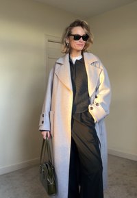 Femme portant des lunettes de soleil noires, un long manteau beige, un pantalon noir, un haut bleu marine, tenant un sac à main vert olive dans une pièce beige avec une porte fermée en arrière-plan.