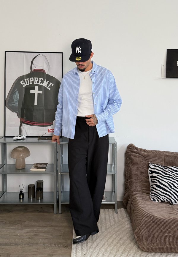 Mees kannab sinise triibulise särgi, valget T-särki, musti laiu pükse ja musta New York Yankees mütsi, seisab minimalistlikus toas koos kunstiteoste ja diivaniga.
