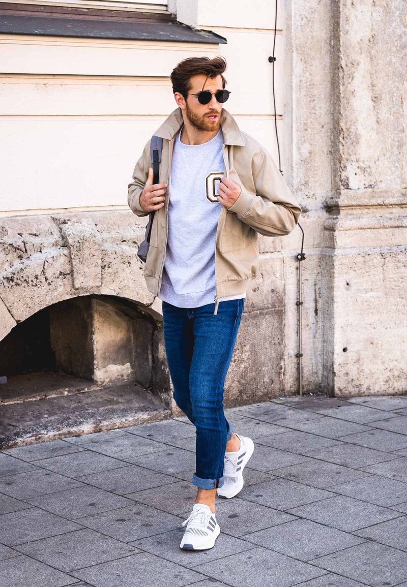 Beige ritsjack over een grijze sweatshirt met een logo, blauwe spijkerbroek en witte sportschoenen met zwarte strepen. Donkere zonnebril gedragen.