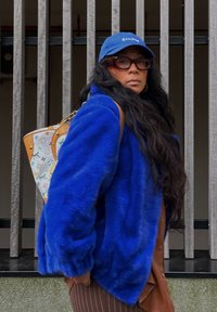 Giacca in finta pelliccia blu con vestibilità rilassata, abbinata a una borsa a tracolla con motivi e loghi multicolori, completata da un cappellino blu.