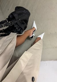 Mules brancos de bico apontado com costas abertas e salto baixo, combinados com um casaco bege. Uma bolsa tecida preta está visível.