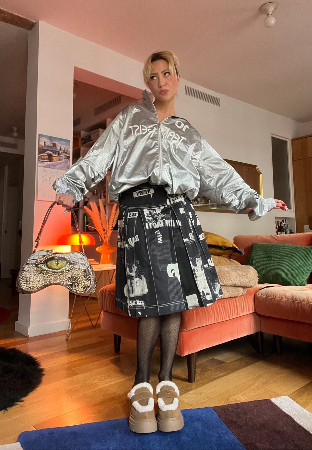 Veste argentée oversize avec logo, jupe plissée noire avec imprimés graphiques, collants texturés, et chaussures compensées marron avec fausse fourrure.