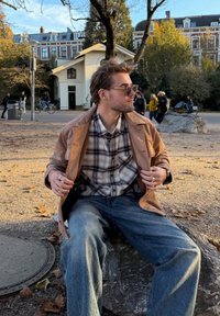 Hnědá kožená bunda přes kostkovanou košili, světle modré džíny, sedící na kameni. Sluneční brýle a viditelné ruce, kolem podzimní listí.