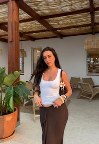 Eine Frau in einem weißen Top und einem braunen Rock steht neben einer Topfpflanze. Sie trägt Accessoires wie Armbänder und hat eine Blume an ihrer Tasche.