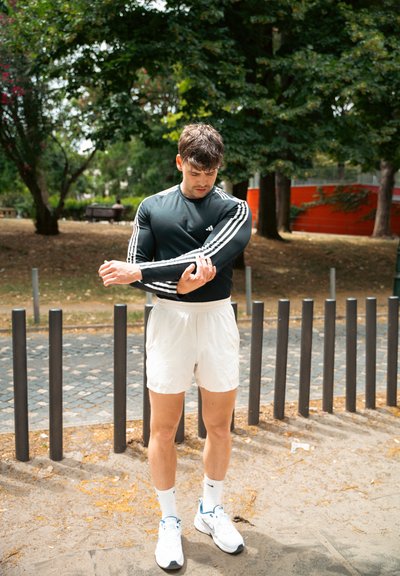 Camisa atlética negra de manga larga con rayas blancas, pantalones cortos blancos y zapatillas blancas. De pie en un parque con árboles y postes de madera al fondo.