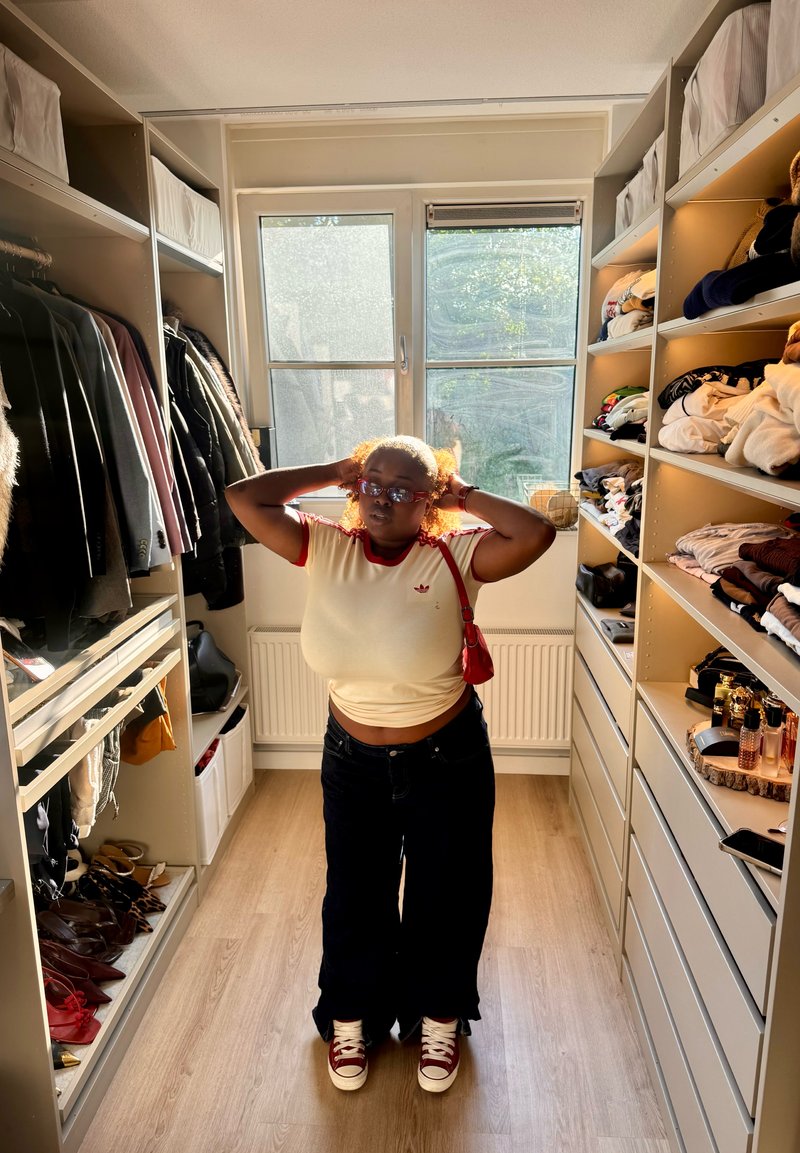 Persoon met krullend haar en bril, die een wit overhemd en een donkere broek draagt, staat in een goed verlichte inloopkast met kleding en schoenen.