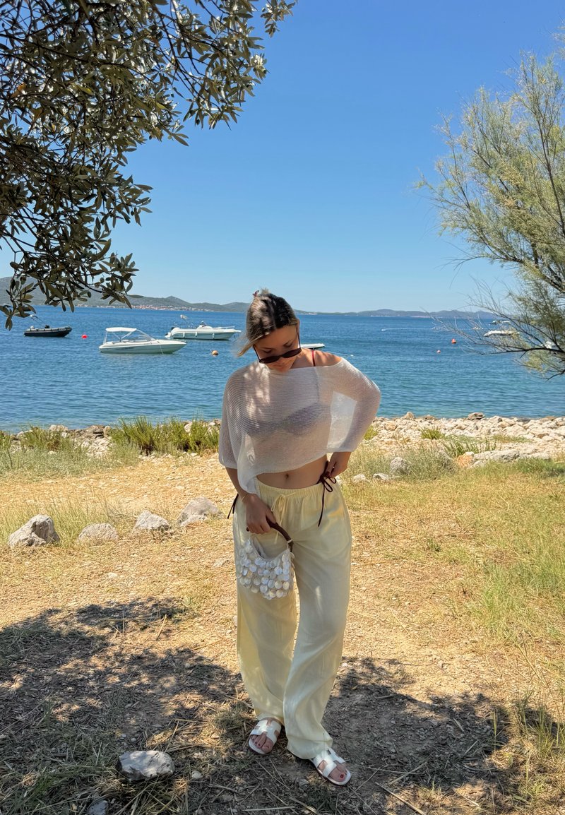 Femme vêtue de vêtements clairs et de lunettes de soleil debout sur l'herbe près d'un rivage rocheux avec des bateaux sur une eau bleue calme sous un ciel dégagé.