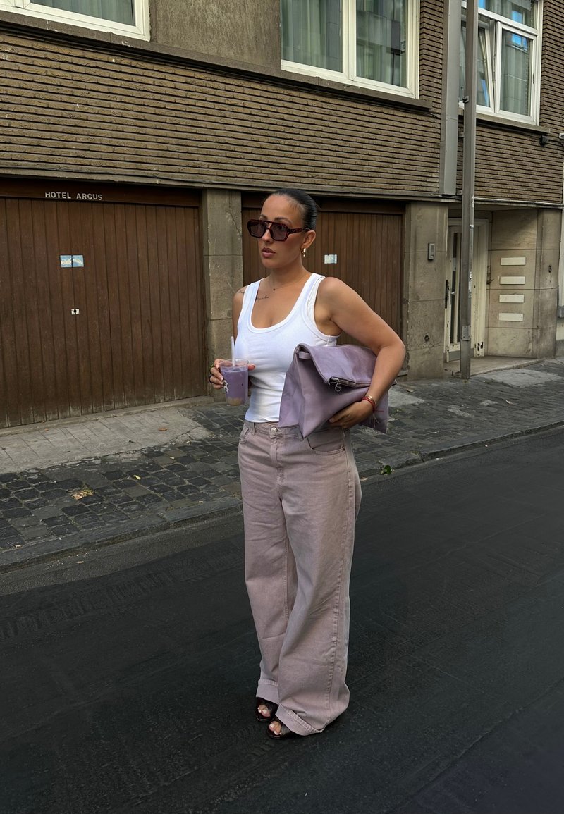 Frau in einem weißen Tanktop und weiten beigen Hosen, die eine große lila Clutch und ein Getränk hält, steht auf einer Straße mit braunen Garagentoren.