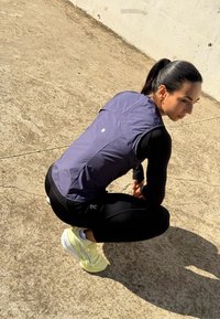 Fioletowa bezrękawnik z wysokim kołnierzem, noszona na czarnym topie z długim rękawem, w połączeniu z czarnymi legginsami i jasnosłomianymi butami do biegania.