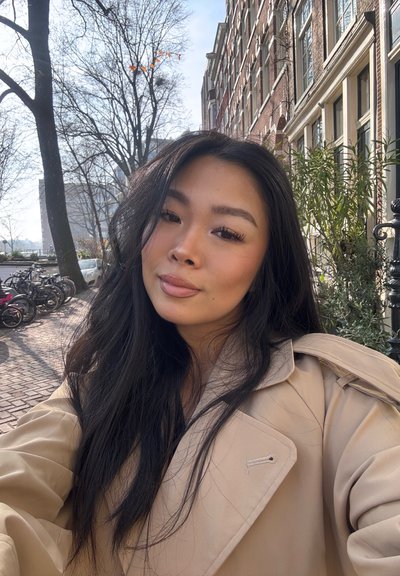 Abrigo trench beige con un ajuste ligeramente oversize, amplio cuello y cierre de gancho único; cabello largo y oscuro; la luz del sol ilumina la escena.