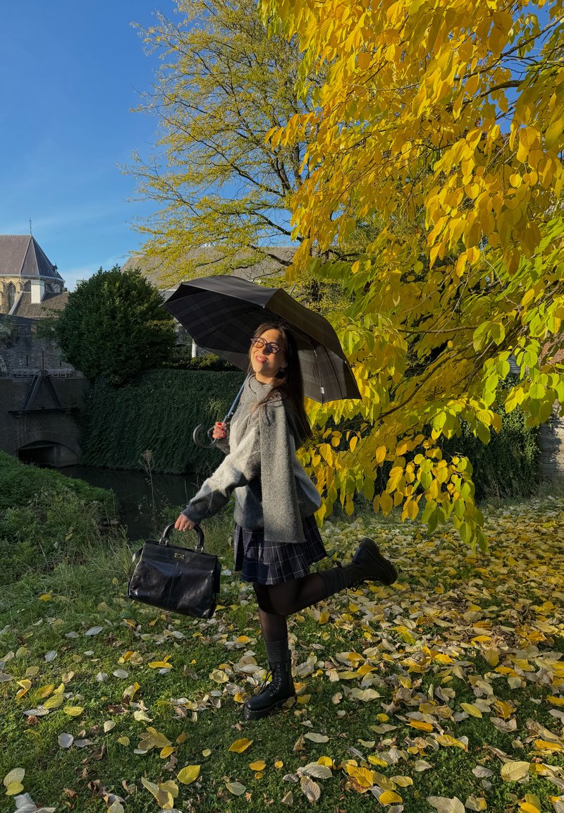 Grau flauschiger Pullover, karierten Rock, schwarze Tasche und schnürbare Stiefel. Hält einen schwarzen Regenschirm gegen gelbe Herbstblätter und den blauen Himmel.