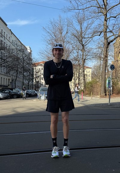 Joven con ropa deportiva negra y zapatillas blancas, de pie con los brazos cruzados en una calle de la ciudad, con árboles sin hojas y edificios al fondo.