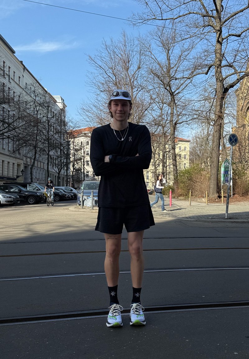 Ung person i sort sportstøj og hvide sneakers står med armene over kors på en bygade med nøgne træer og bygninger i baggrunden.