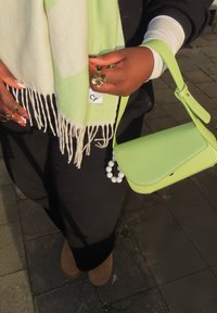 Χέρια με δαχτυλίδια από λουλούδια και χρυσό κρατούν ένα ανοιχτό πράσινο κασκόλ με φούντες και μια πράσινη τσάντα που ταιριάζει, πάνω από μαύρο παντελόνι και καφέ παπούτσια.