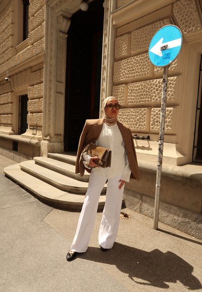 Blazer marrón, blusa blanca y pantalones de pierna ancha blancos. Los accesorios incluyen un bolso dorado. Las gafas de sol y el hijab completan el look. Escena callejera.