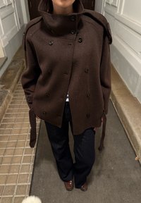 Brązowa dwurzędowa kurtka z wysokim kołnierzem, wykonana z miękkiej tkaniny, z ozdobnymi guzikami i detalami wiązania na rękawach.