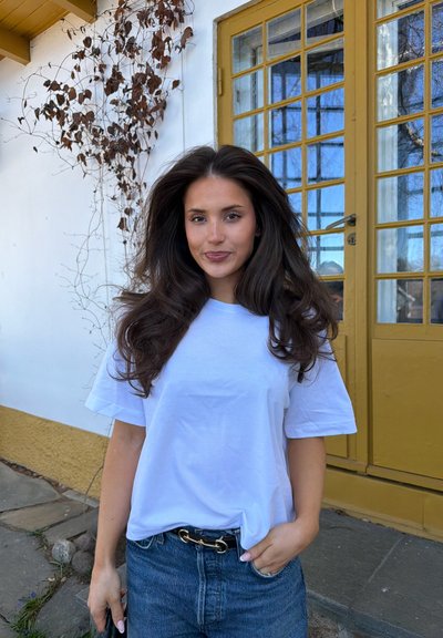 Joven mujer con cabello largo y oscuro, que lleva una camiseta blanca y vaqueros azules, está cerca de una puerta color mostaza con enredaderas sin hojas en una pared blanca.