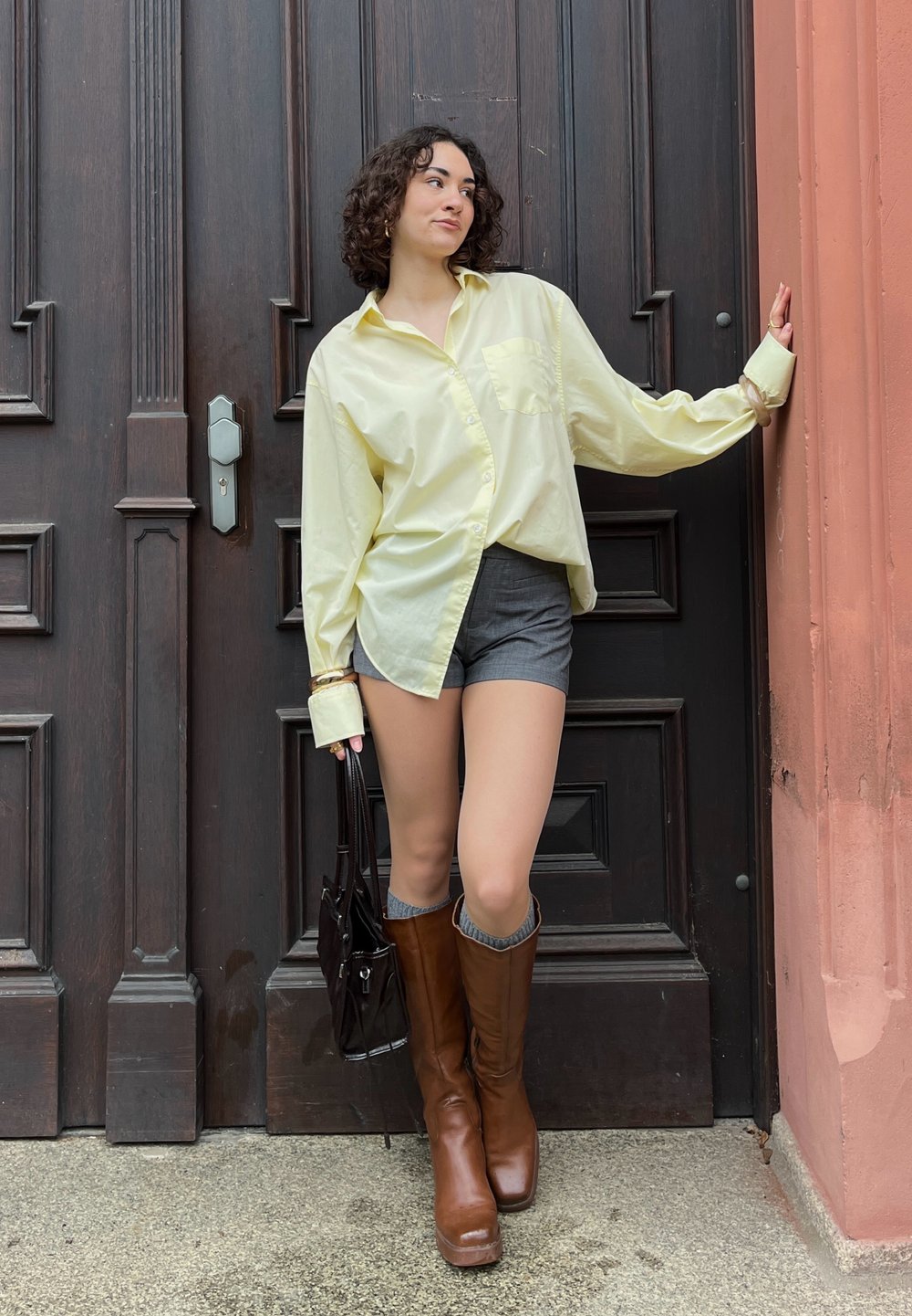 Camisa solta de botão amarelo claro, calções cinzentos, botas de couro castanho até ao joelho e uma mala escura. Posada de forma estilosa contra uma porta de madeira.