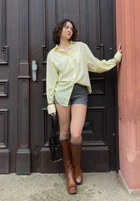 Camisa solta de botão amarelo claro, calções cinzentos, botas de couro castanho até ao joelho e uma mala escura. Posada de forma estilosa contra uma porta de madeira.