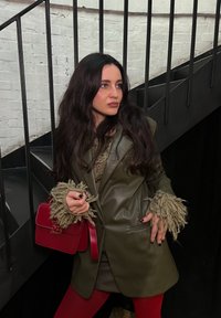 Blazer de cuero verde oliva con mangas texturizadas y flecos, combinado con un bolso rojo y leggins burdeos, contra unas escaleras negras.